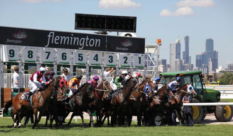 Horses burst from the starting gates at Flemington Racecourse in Melbourne, with jockeys in colourful silks and the city skyline in the background.