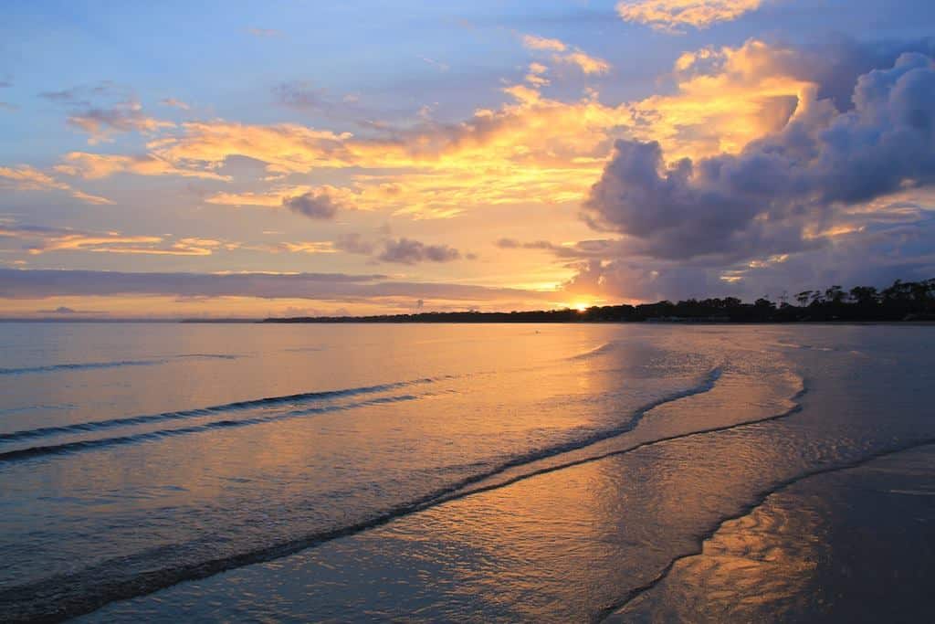 The sun rises over a calm bay through a mountain of cloud. # wide ripples roll toward shore and the sun's light is reflected on the water.