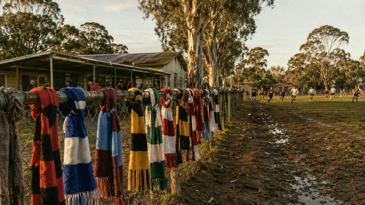 Colourful vintage soccer scarves hang on a wire fence at a muddy suburban Australian soccer ground, with gum trees, an old clubhouse, spectators, and a match in progress, evoking the migrant roots of Australian football.